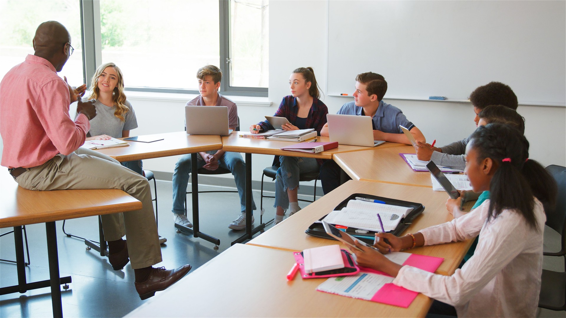 Male High School Tutor Sitting On Desk And Asking Students With Laptops And Digital Tablets Question